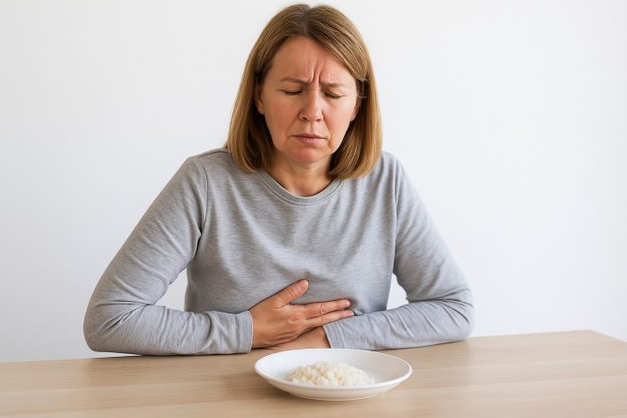 Femeie de vârstă mijlocie stând la masă, ținându-se de abdomenul superior și privind cu disconfort spre farfuria neatinsă – sugestie vizuală pentru simptomele cancerului gastric.
