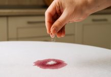 Trucul cu sare care face minuni: cum salvezi hainele pătate fără detergent. Femeile îl adoră! Mână care presară sare peste o pată de vin roșu pe o față de masă albă