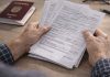 Românii care au lucrat în străinătate: ce drepturi au la pensie și ce trebuie să știe în 2026 Mâinile unei persoane adulte ținând documente de muncă și formulare, lângă un pașaport românesc deschis, pe o masă, într-un context de verificare și decizie