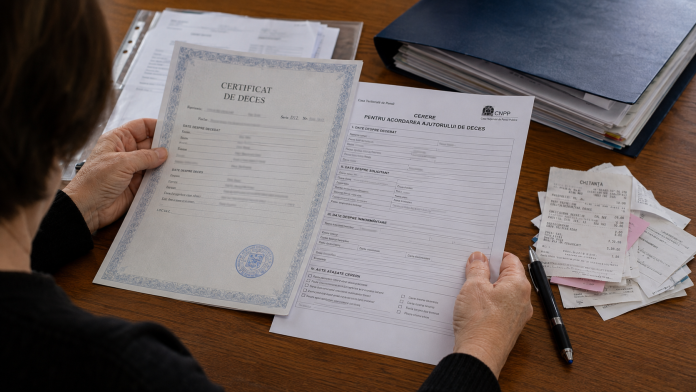 ajutor-de-deces-2026.jpg Mâinile unei persoane analizând documente pentru acordarea ajutorului de deces pe un birou.