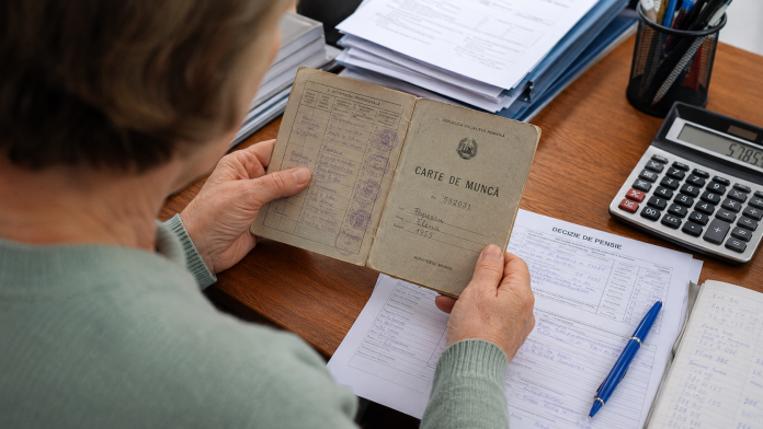 vechime-pensie-documente.jpg Persoană analizând carnet de muncă și documente de pensie pe un birou.