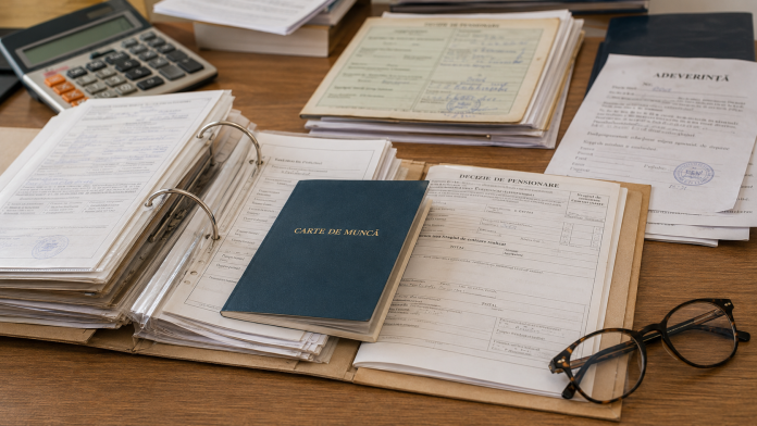 verificare-stagiu-cotizare.jpg Dosar de pensionare deschis, carte de muncă, adeverințe și documente privind stagiul de cotizare analizate pe un birou.